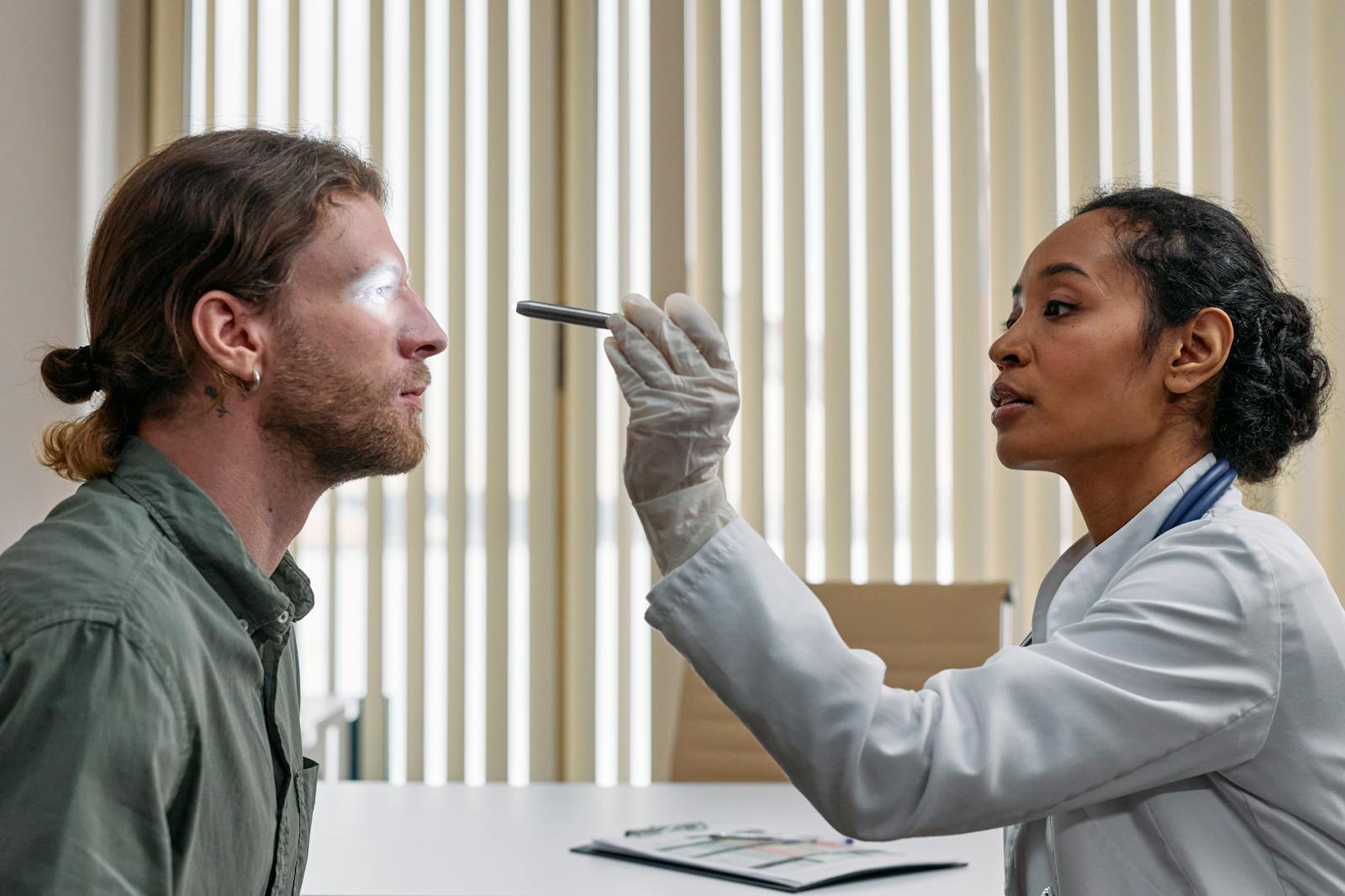 A doctor performs an eye examination on a patient using a flashlight in a clinical setting.