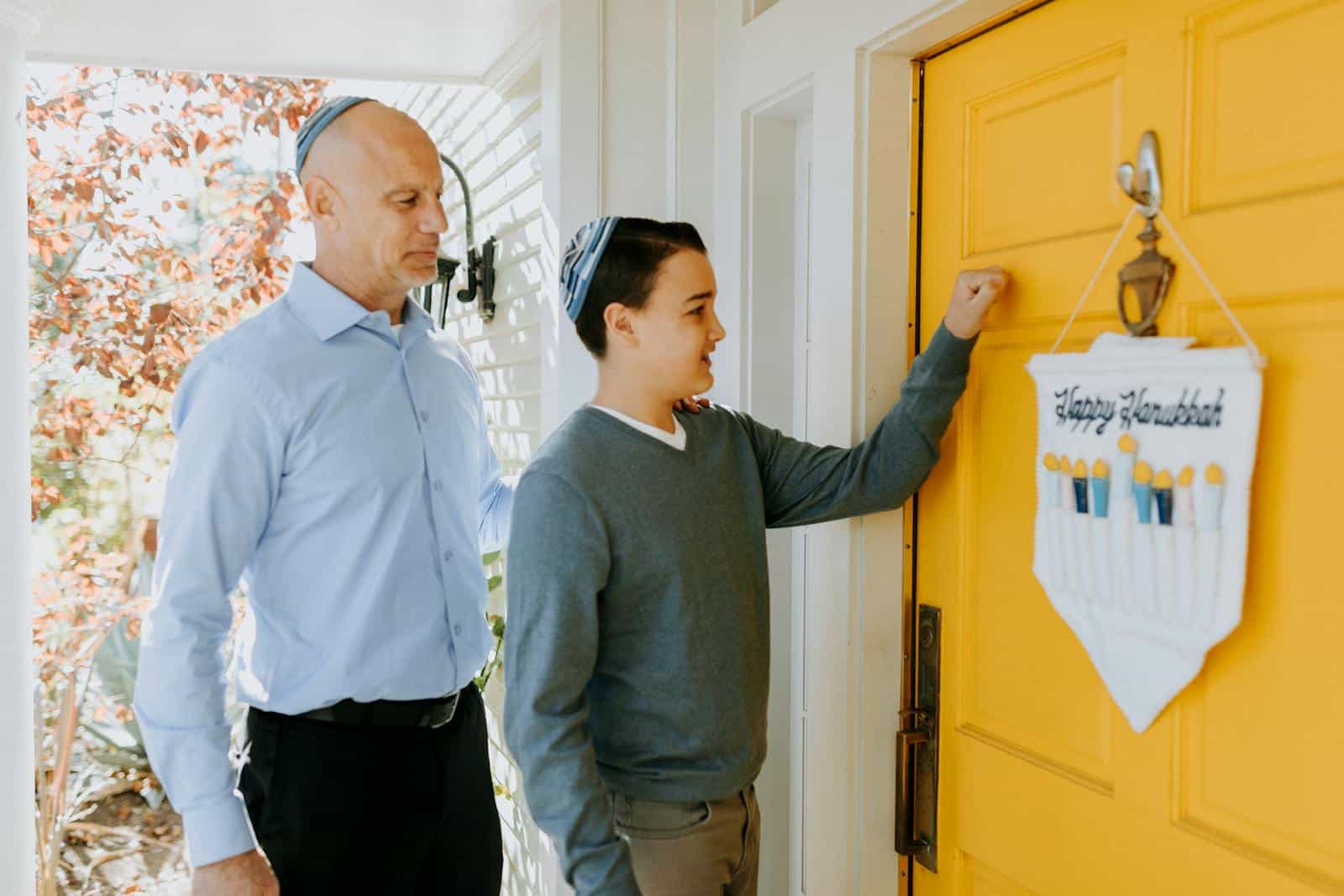 A father and son knocking on a yellow door with a Happy Hanukkah sign, celebrating the Jewish holiday.
