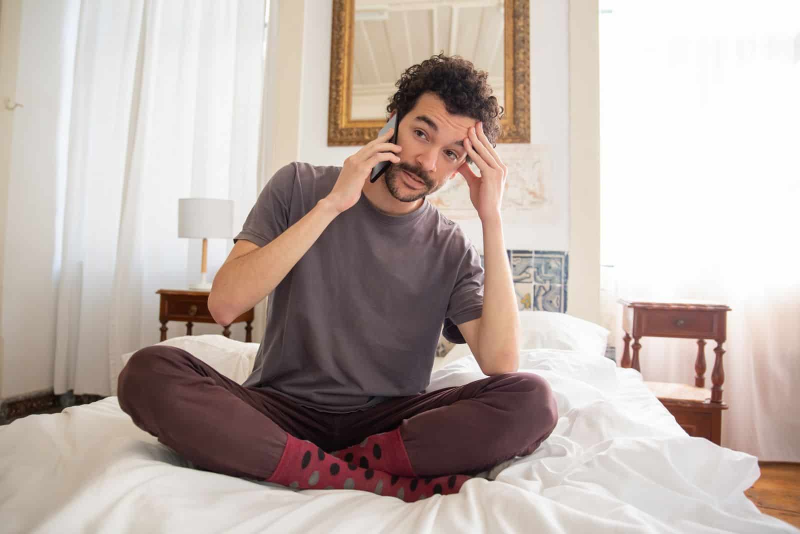 A man in casual attire sits on a bed talking on the phone with a thoughtful expression.