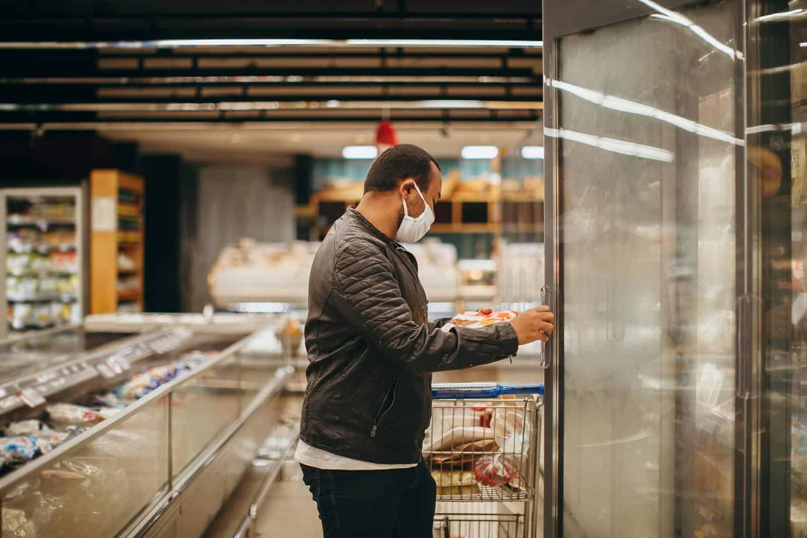 A man wearing a mask shops in a supermarket, selecting items from a refrigerated section. grocery shopping
