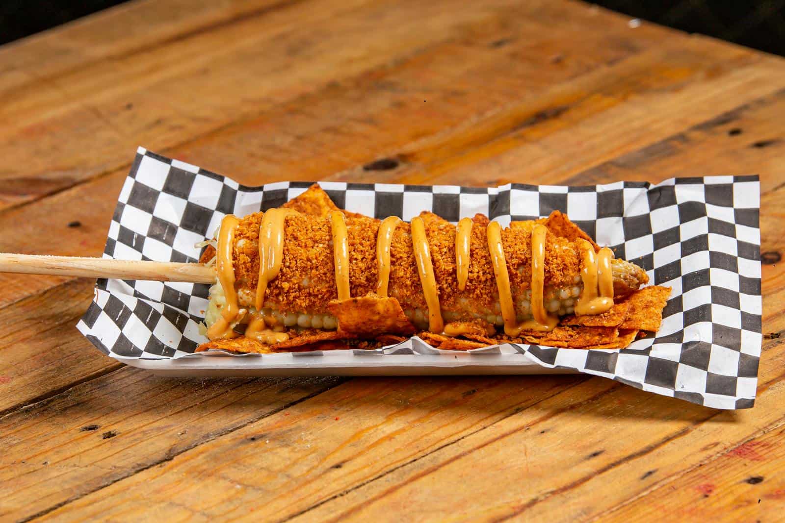 A mouth-watering fried corn on a stick drizzled with sauce, served on a checkered paper.