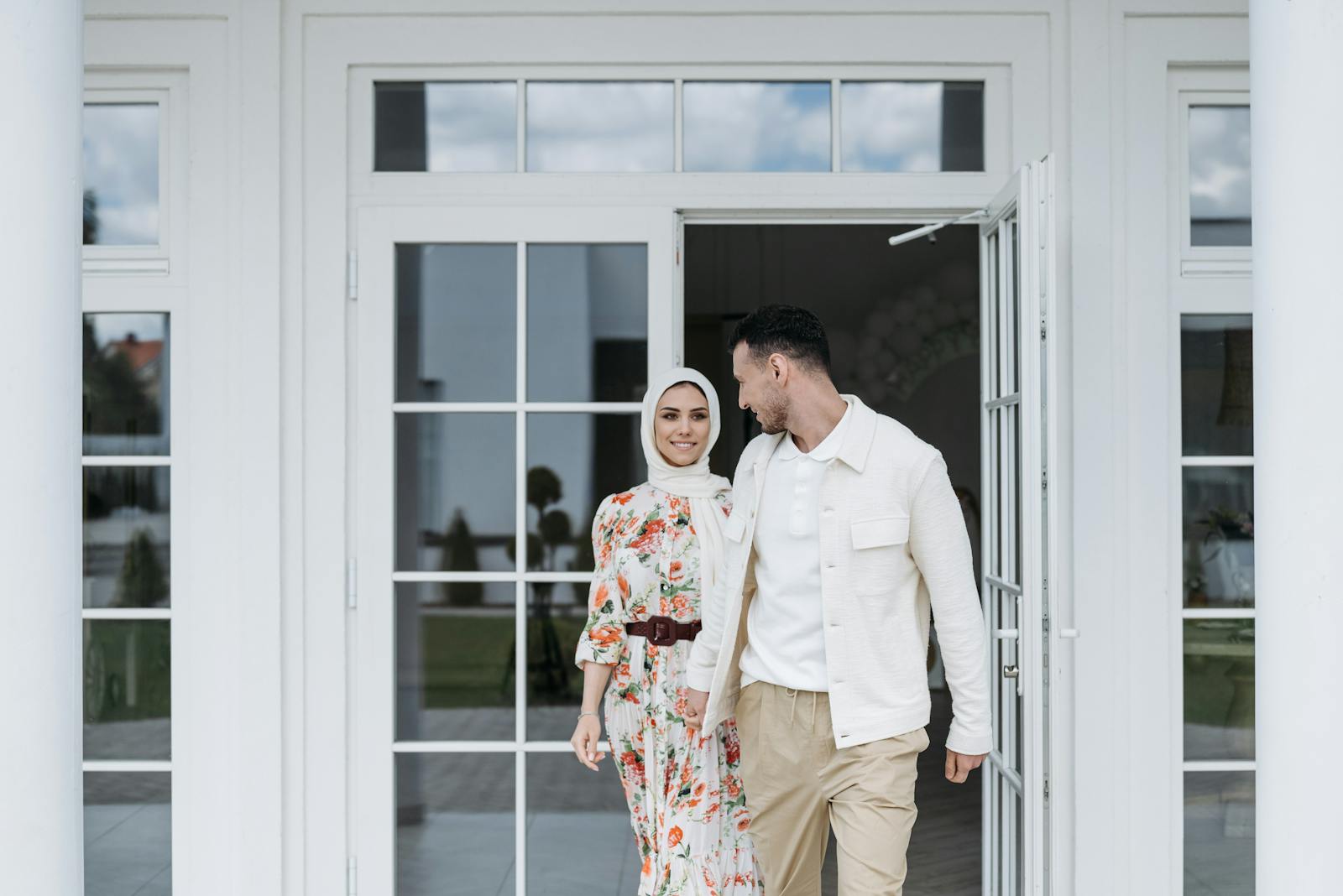 A smiling couple stands in a modern doorway with glass windows, featuring a floral dress and outerwear.