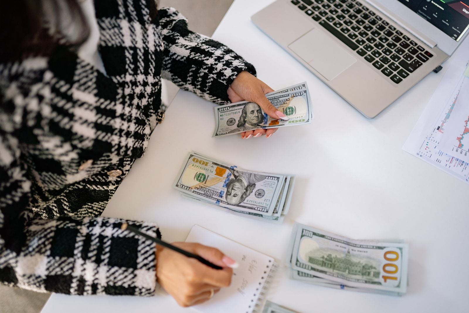 A woman in a plaid coat counts American dollars at a desk with a notebook and laptop.
