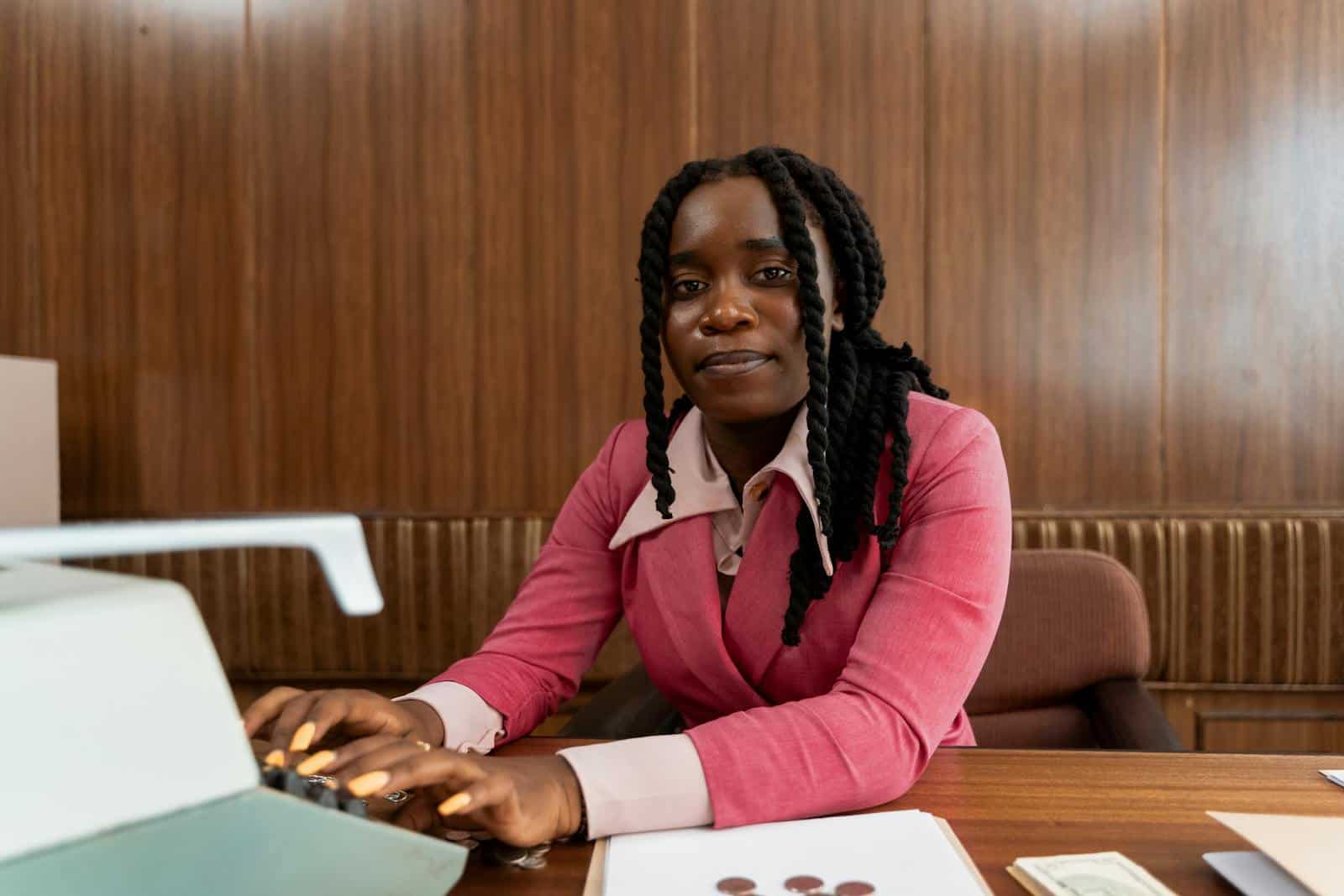 African American woman in a pink suit typing on a retro typewriter in an office setting.