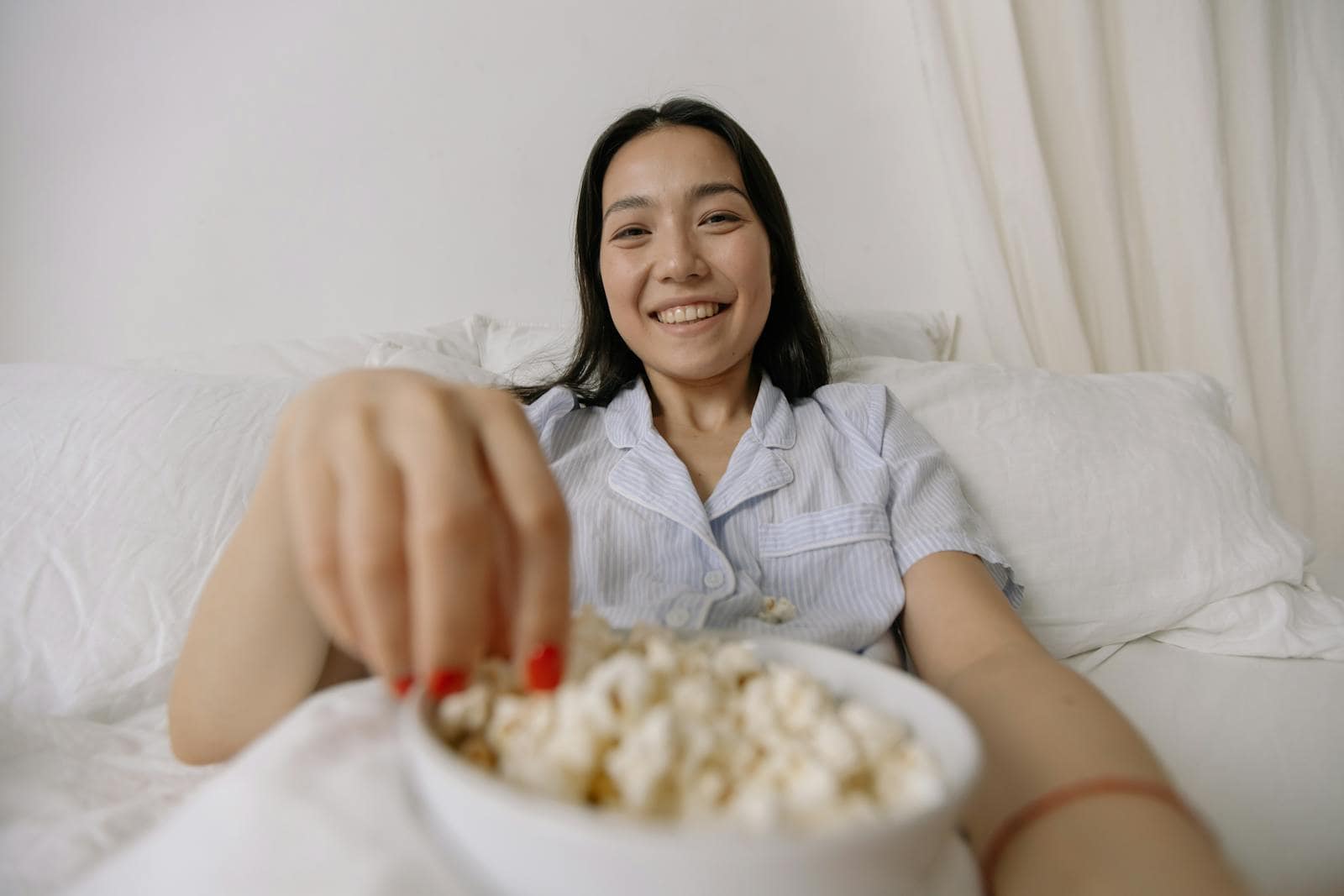 Asian woman in pajamas smiling while eating popcorn in a cozy bedroom setting.