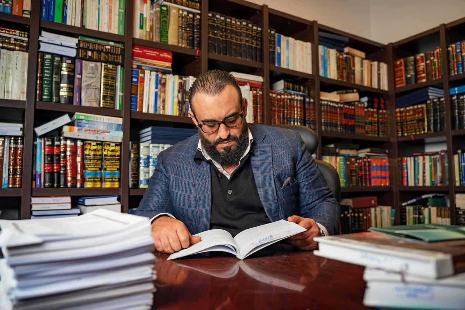 Bearded man reading a book in a library, wearing eyeglasses and a blue suit. Ideal for education themes.