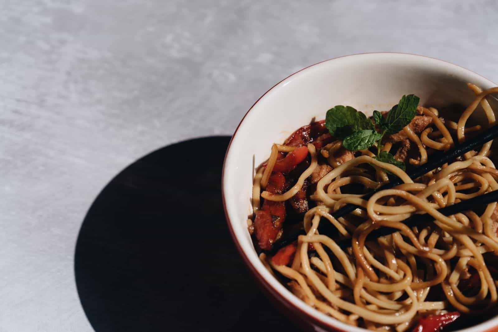Bowl of Asian noodles with black chopsticks, seasoned vegetables, and fresh mint garnish.