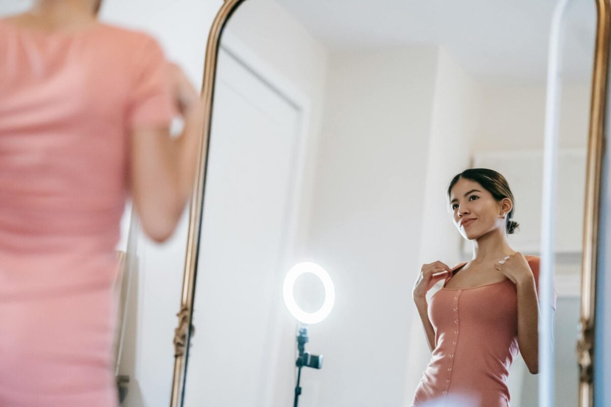 Content young female blogger in pink dress looking in mirror against illuminating ring lamp in light room