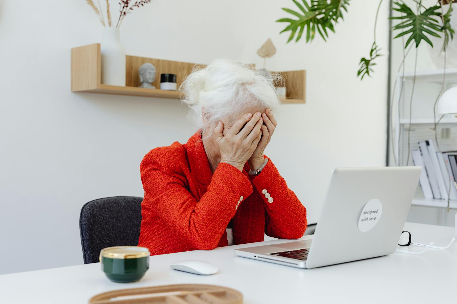 Elderly woman in red jacket looking frustrated at laptop in home office setting.