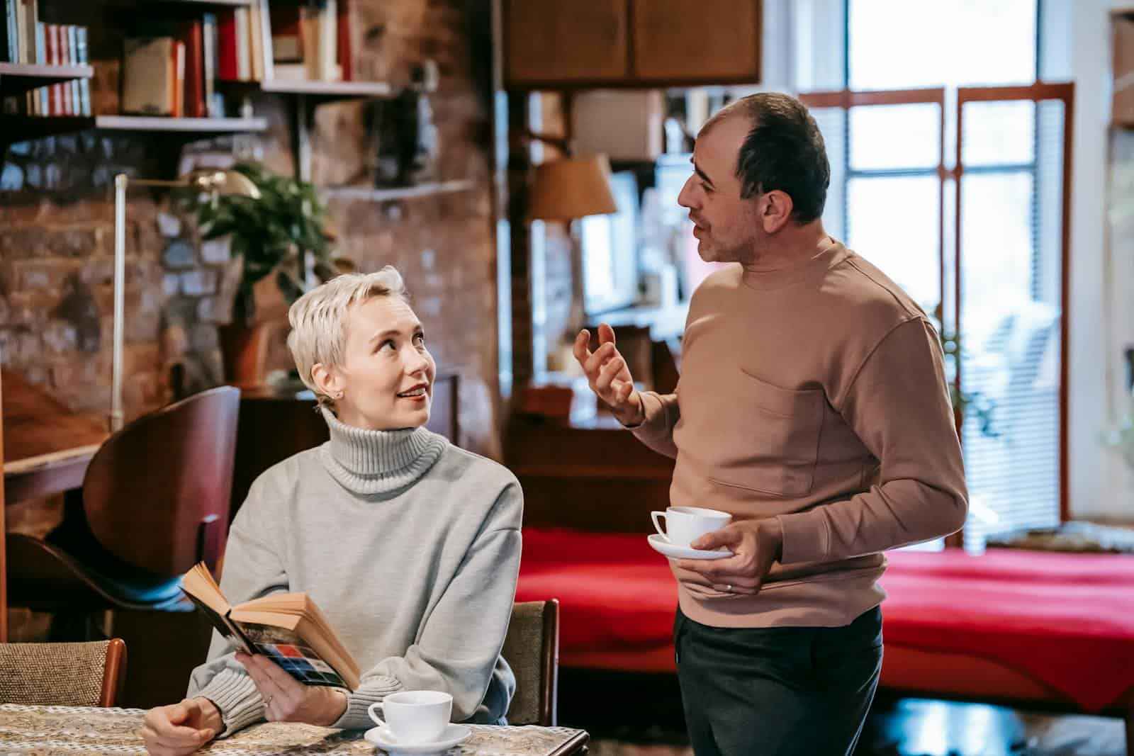 Positive diverse middle aged spouses in casual clothes drinking coffee and discussing interesting book while spending weekend together at home senior