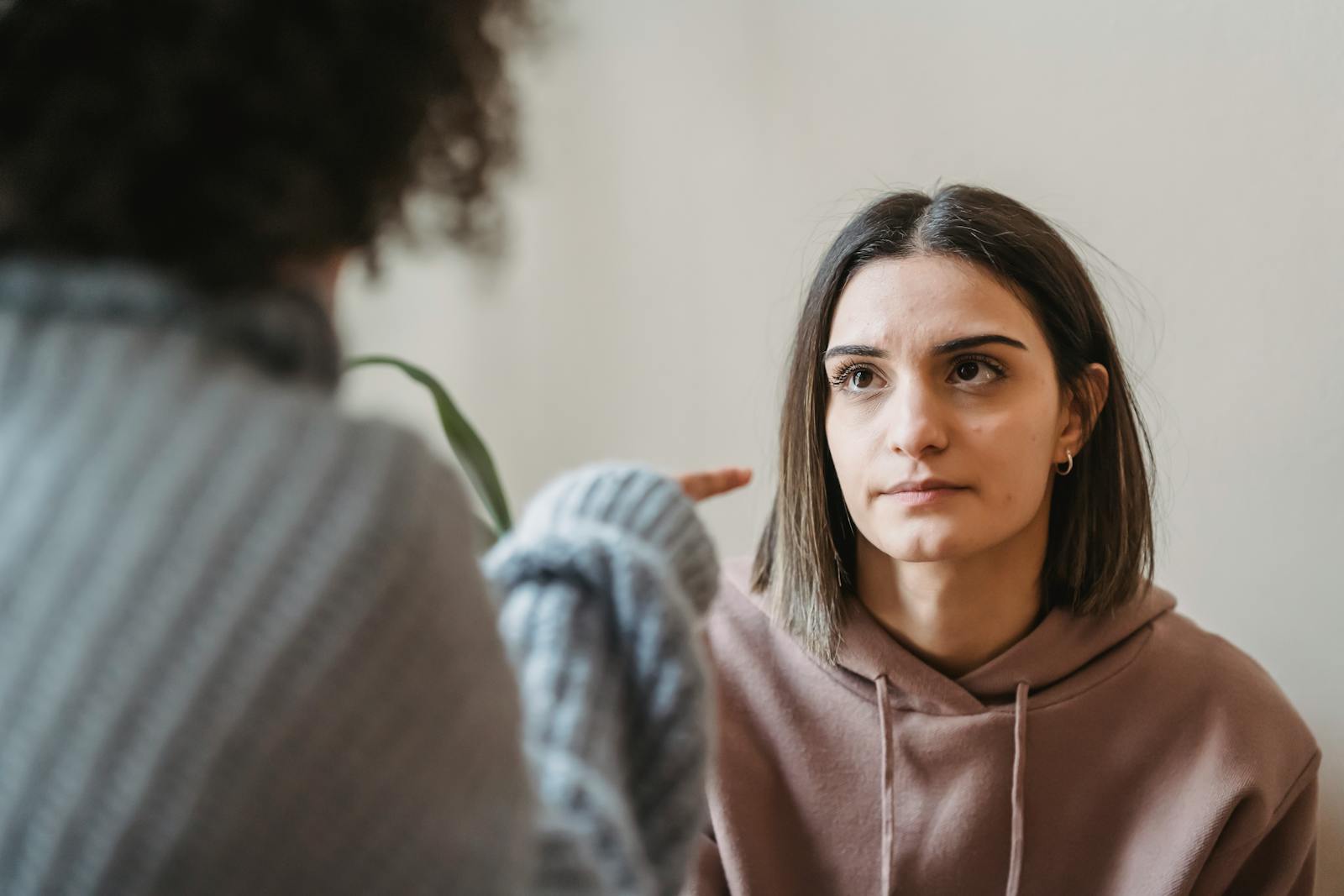 Sad young ladies in casual clothes standing in light apartment near wall while having disagreement and pointing finger in face while looking at each other