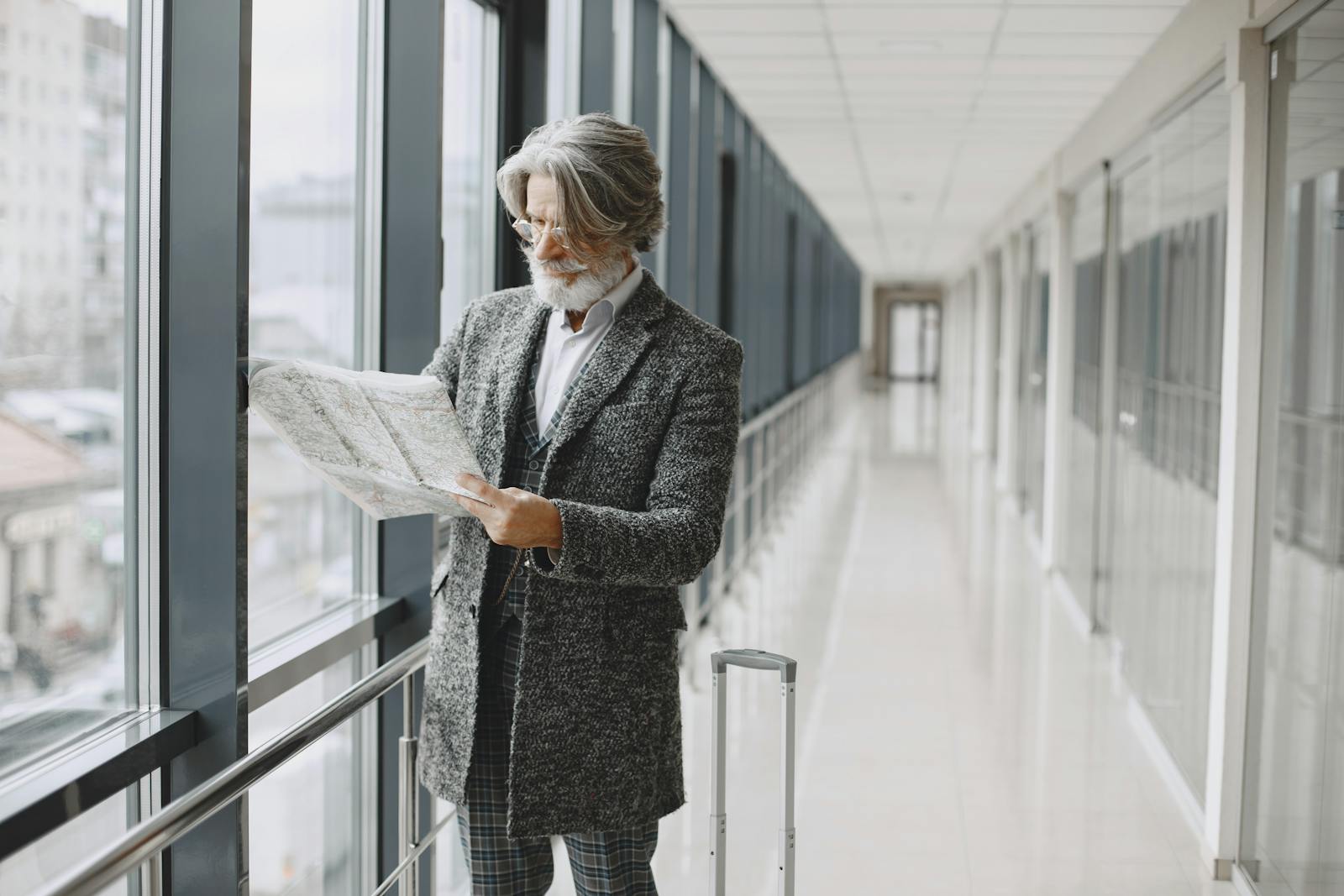 Senior man with beard reads map by glass windows in modern hallway with luggage.business trip
