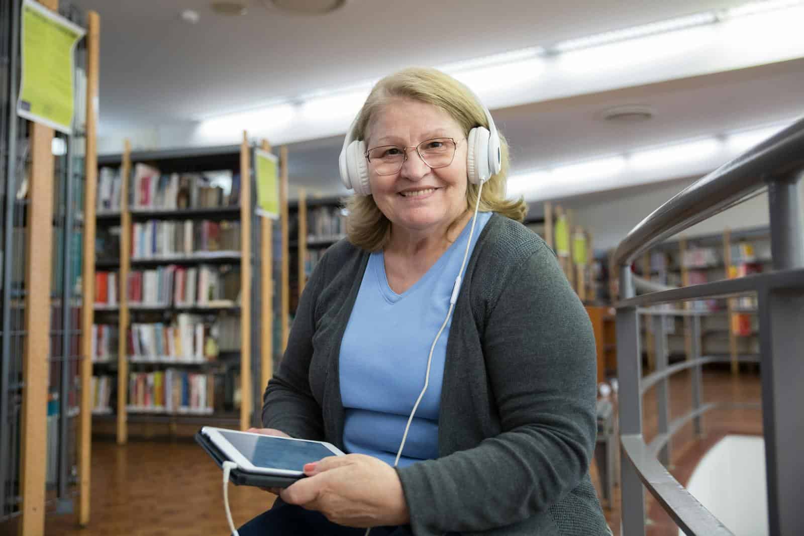 Senior woman smiling while listening to audiobooks on tablet in library setting. audiobook