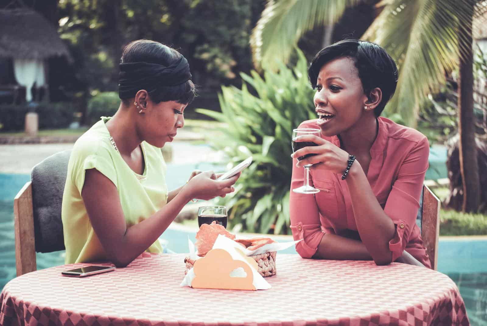Two women enjoying a relaxing outdoor lunch by the pool, sipping drinks and chatting.