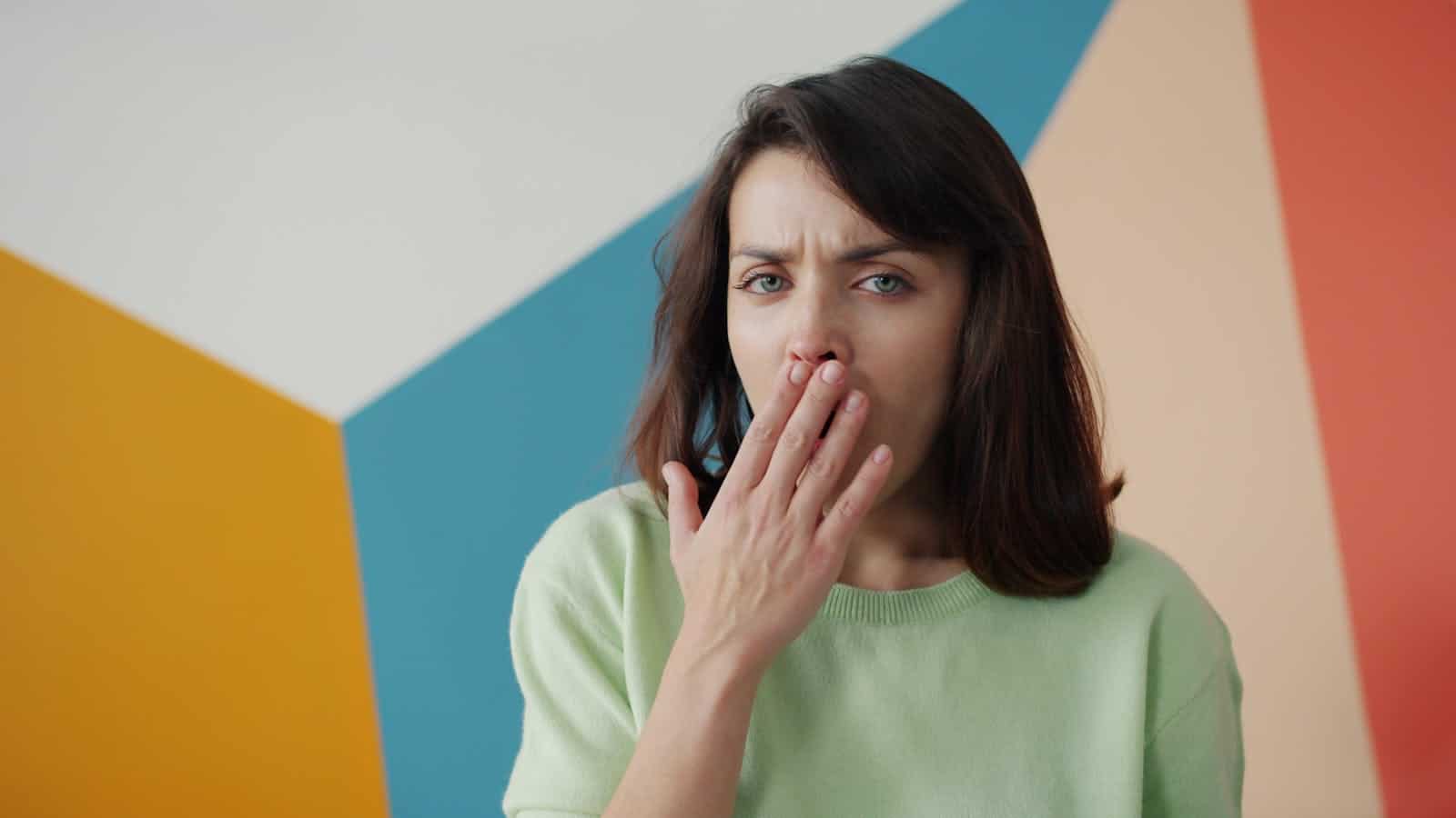 Woman covers her mouth against a colorful geometric wall, expressing surprise or yawn.