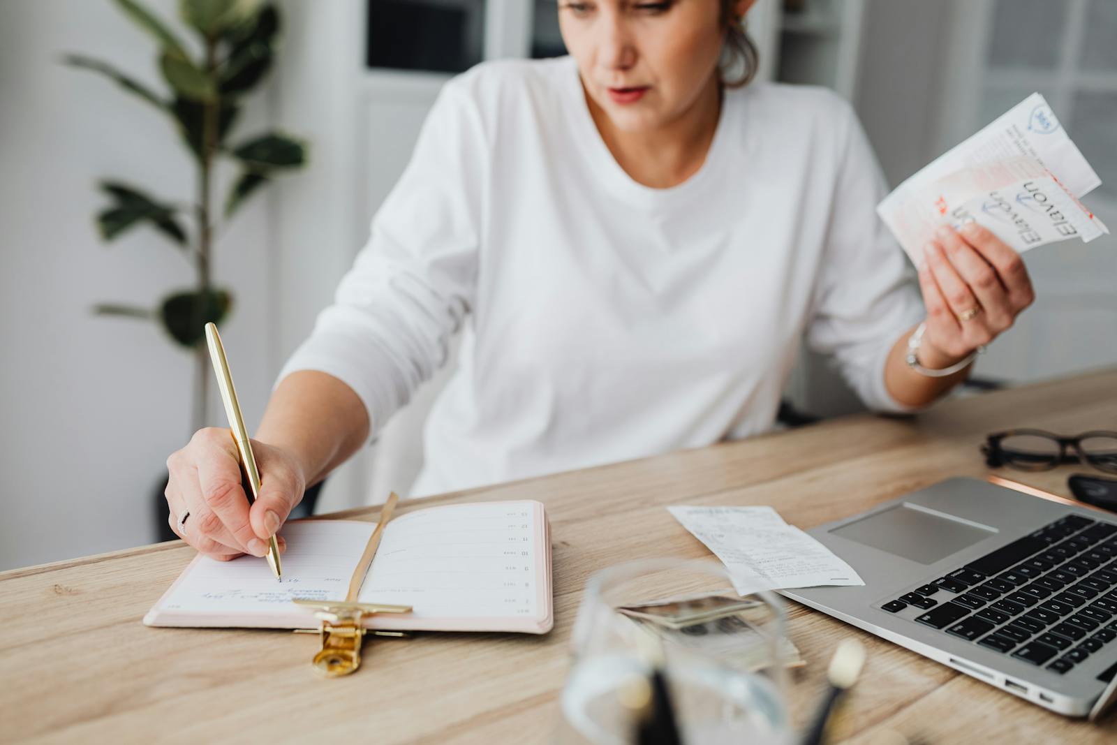 Woman managing finances, writing on notepad, and holding receipts at a desk.