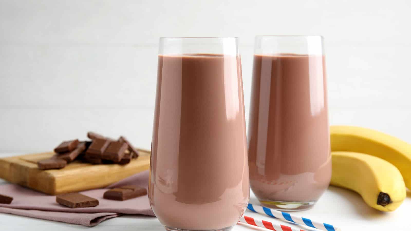 Two glasses of chocolate milkshake with bananas in the background.