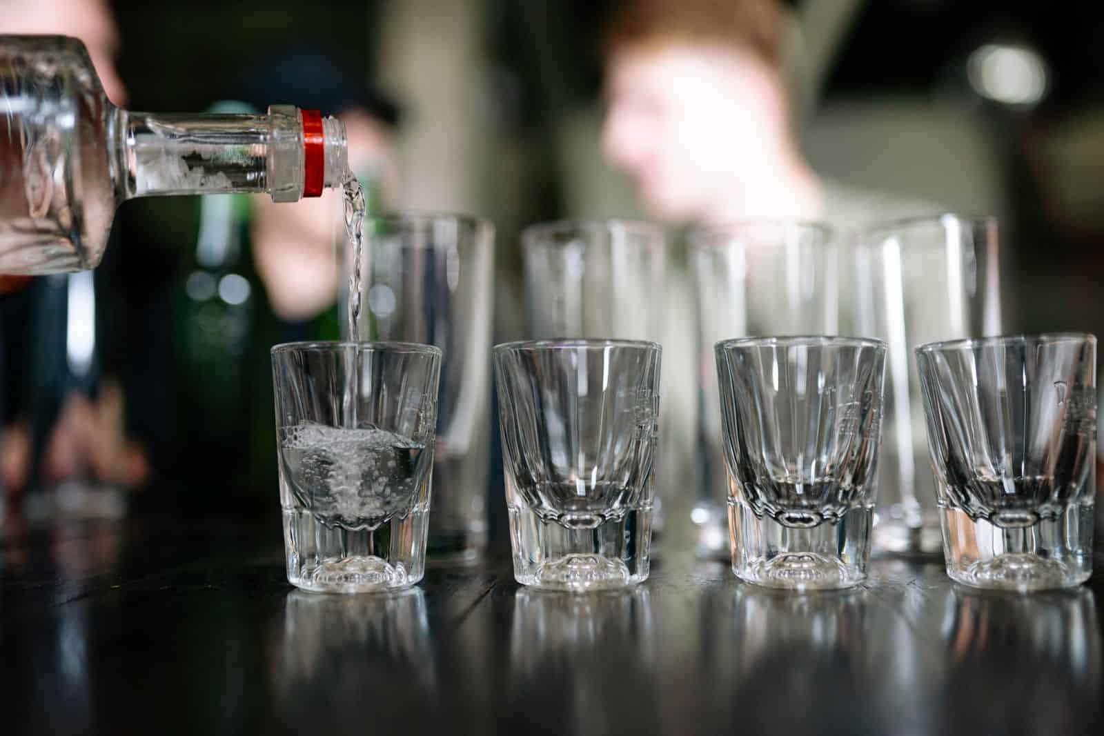 Close-up of a bottle pouring clear alcohol into shot glasses, with people in the background. White Liquor