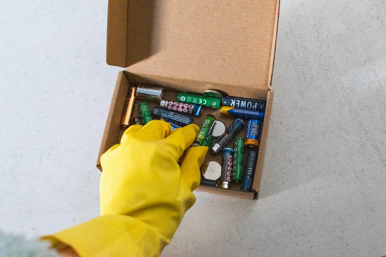 Yellow-gloved hand handling used batteries for safe recycling. Button Batteries