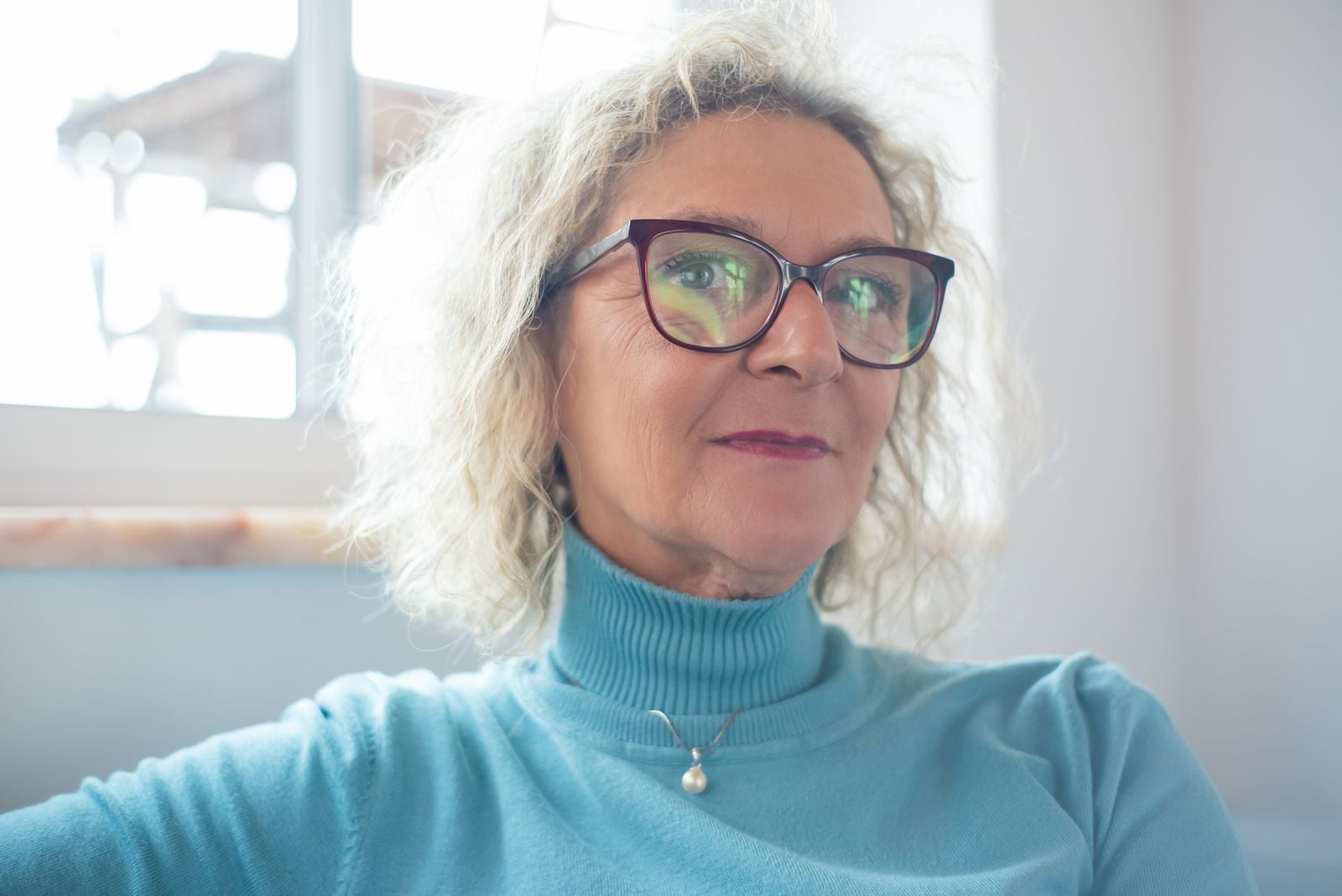 Elderly woman wearing glasses and a turtleneck sweater, sitting indoors with natural light.