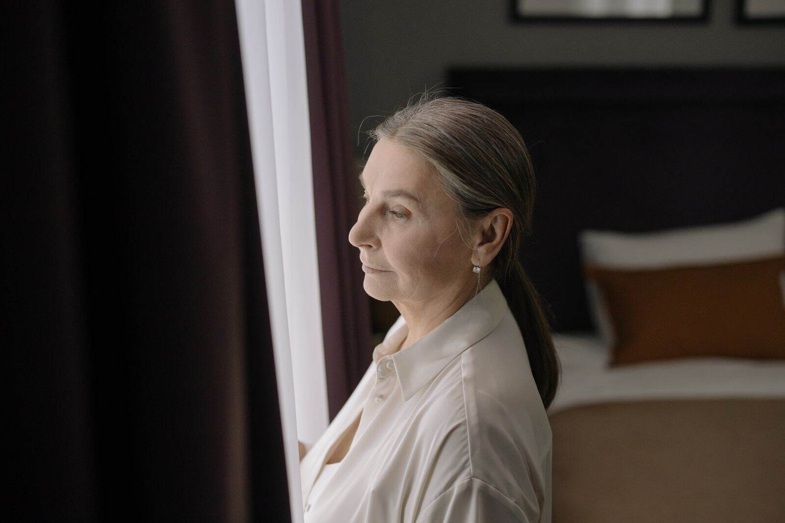Senior woman with grey hair in profile, gazing out a window, conveying peaceful reflection.