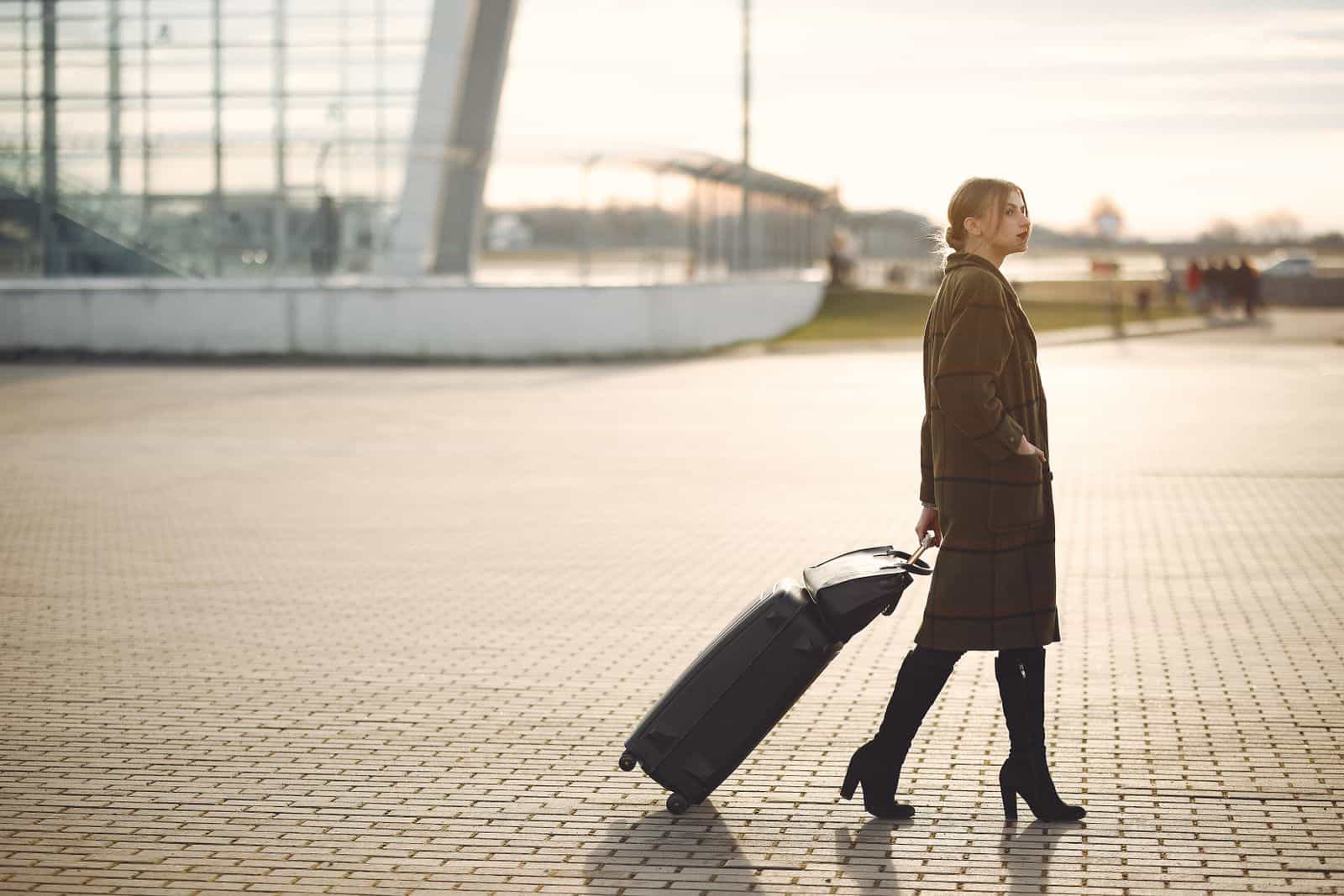 Side view of full length young female traveler in trendy plaid brown coat and high boots with suitcase walking on pavement near modern building on city street in morning Expatriate