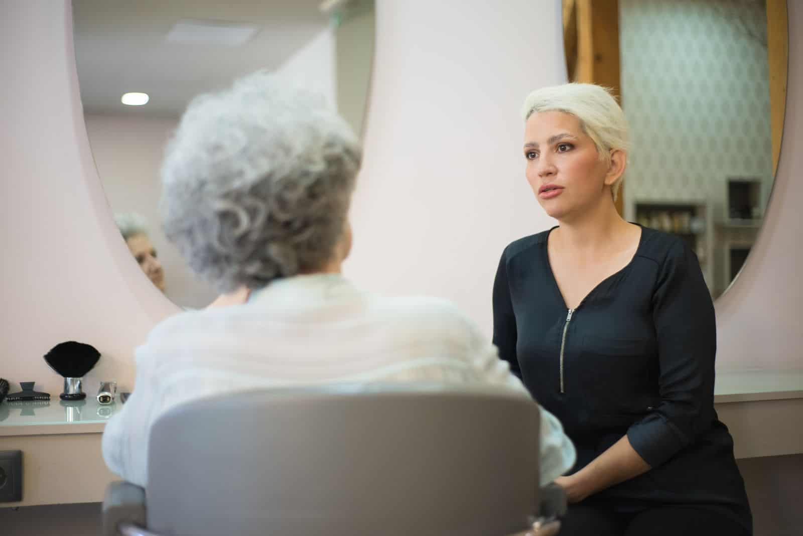 Two women having a conversation in a salon setting with a mirror reflecting their interaction.